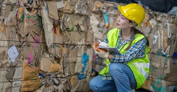 A woman wearing a yellow hard hat and high-visibility vest inspects and takes notes about bales of compressed cardboard at a recycling facility, kneeling beside stacked bundles of flattened boxes secured with wire.