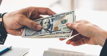 Two hands exchanging hundred dollar bills in cash transaction over desk with papers and pen.