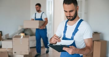 A moving company worker in overalls writing on a clipboard while his colleague carries a cardboard box in the background, surrounded by packed boxes in a bright residential room during a relocation service.