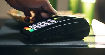 Hand holding credit card over contactless payment terminal for tap-to-pay transaction at retail counter.