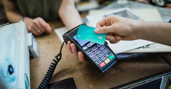 Hand holding teal credit card over payment terminal for contactless transaction at retail counter.