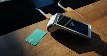A contactless payment terminal and teal credit card sit on a wooden table with wireless payment symbols visible.