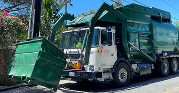 A green and white garbage truck with hydraulic lifting arms is collecting waste from large green dumpsters on a residential street lined with tropical vegetation under a clear blue sky.