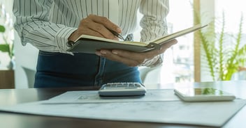 A person in a striped shirt writes in a notebook while standing at a desk with a calculator and papers nearby.