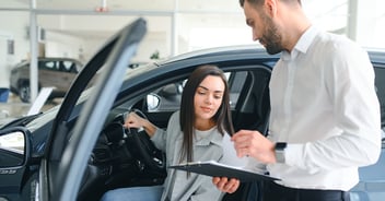 A female client stepping into a car while looking over details on a clipboard with an auto dealer.