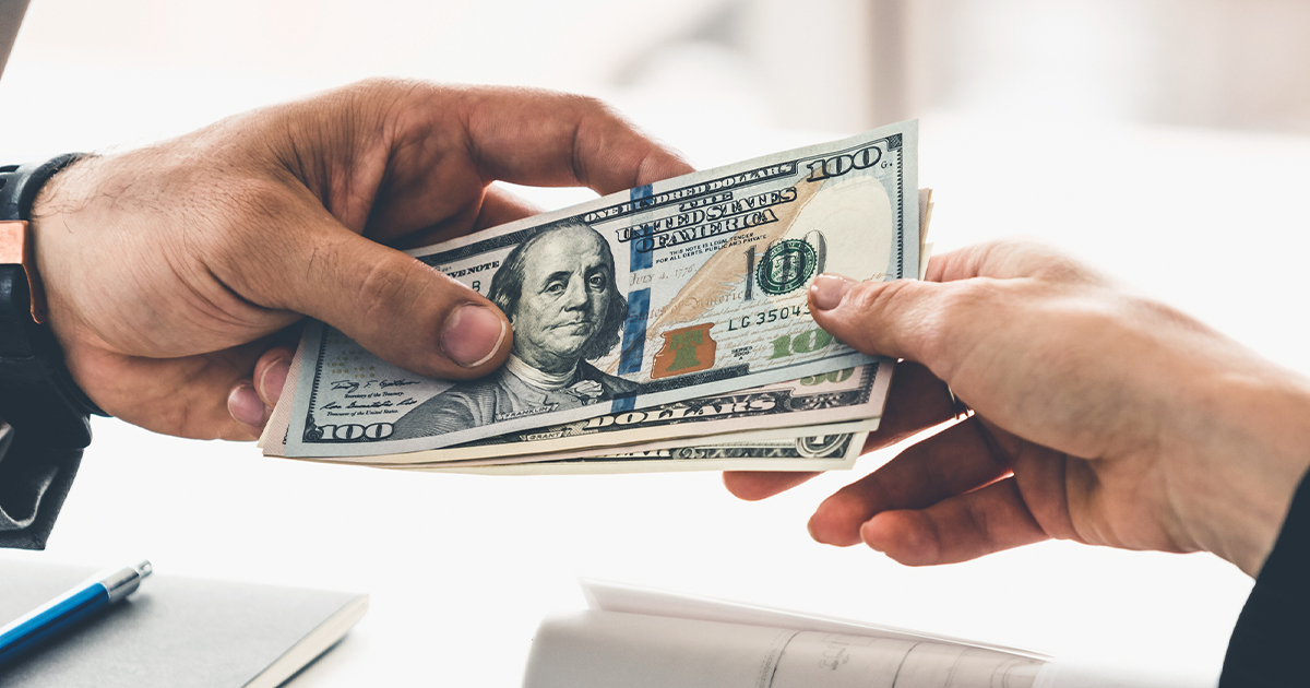 Two hands exchanging hundred dollar bills in cash transaction over desk with papers and pen.