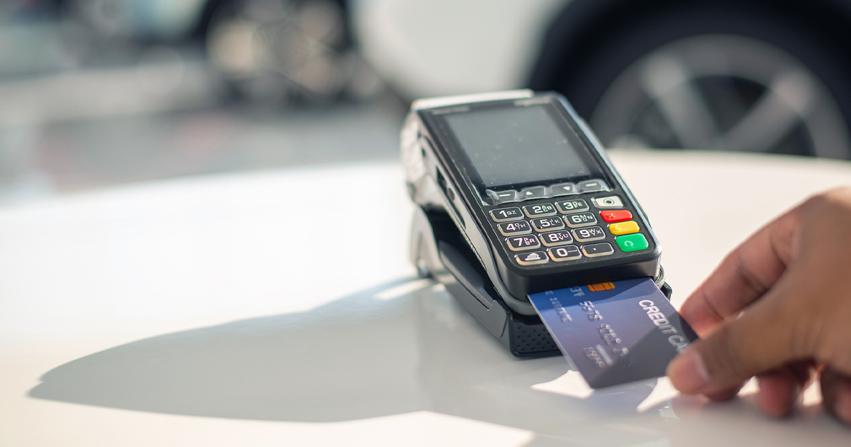 A person's hand inserting a blue credit card into a point-of-sale terminal with a numeric keypad and colored function buttons on a white counter surface.
