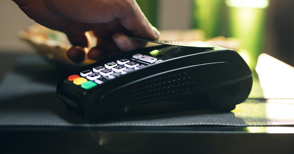 Hand holding credit card over contactless payment terminal for tap-to-pay transaction at retail counter.