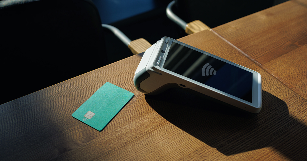A contactless payment terminal and teal credit card sit on a wooden table with wireless payment symbols visible.