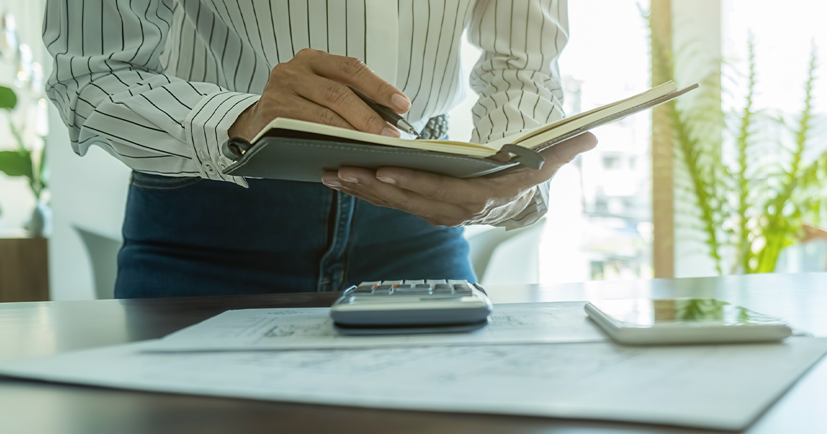 A person in a striped shirt writes in a notebook while standing at a desk with a calculator and papers nearby.